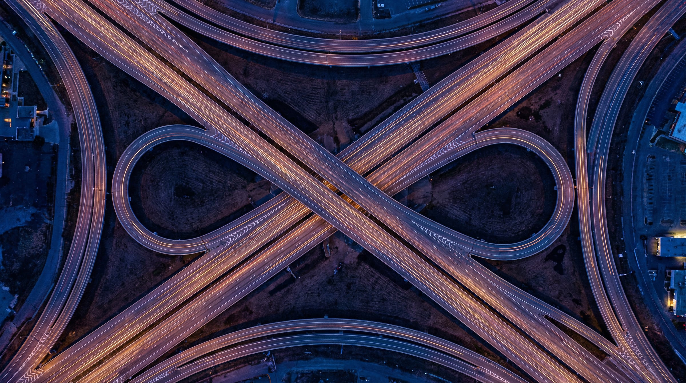 Aerial view at dusk of a highway interchange, with light trails tracing its looping ramps.